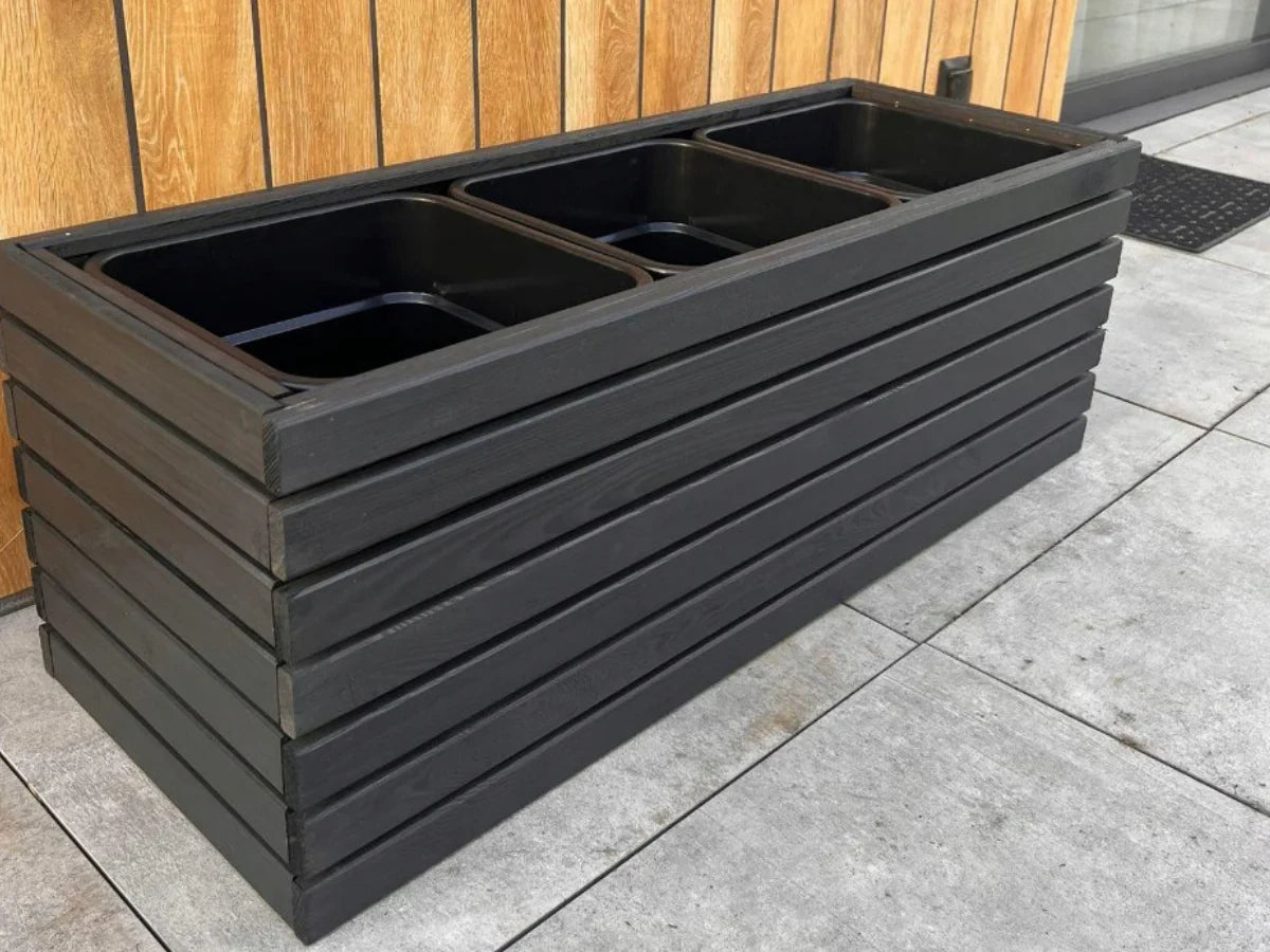 Czarna donica drewniana z wkładami, ustawiona przy ścianie na tarasie z płytami betonowymi.